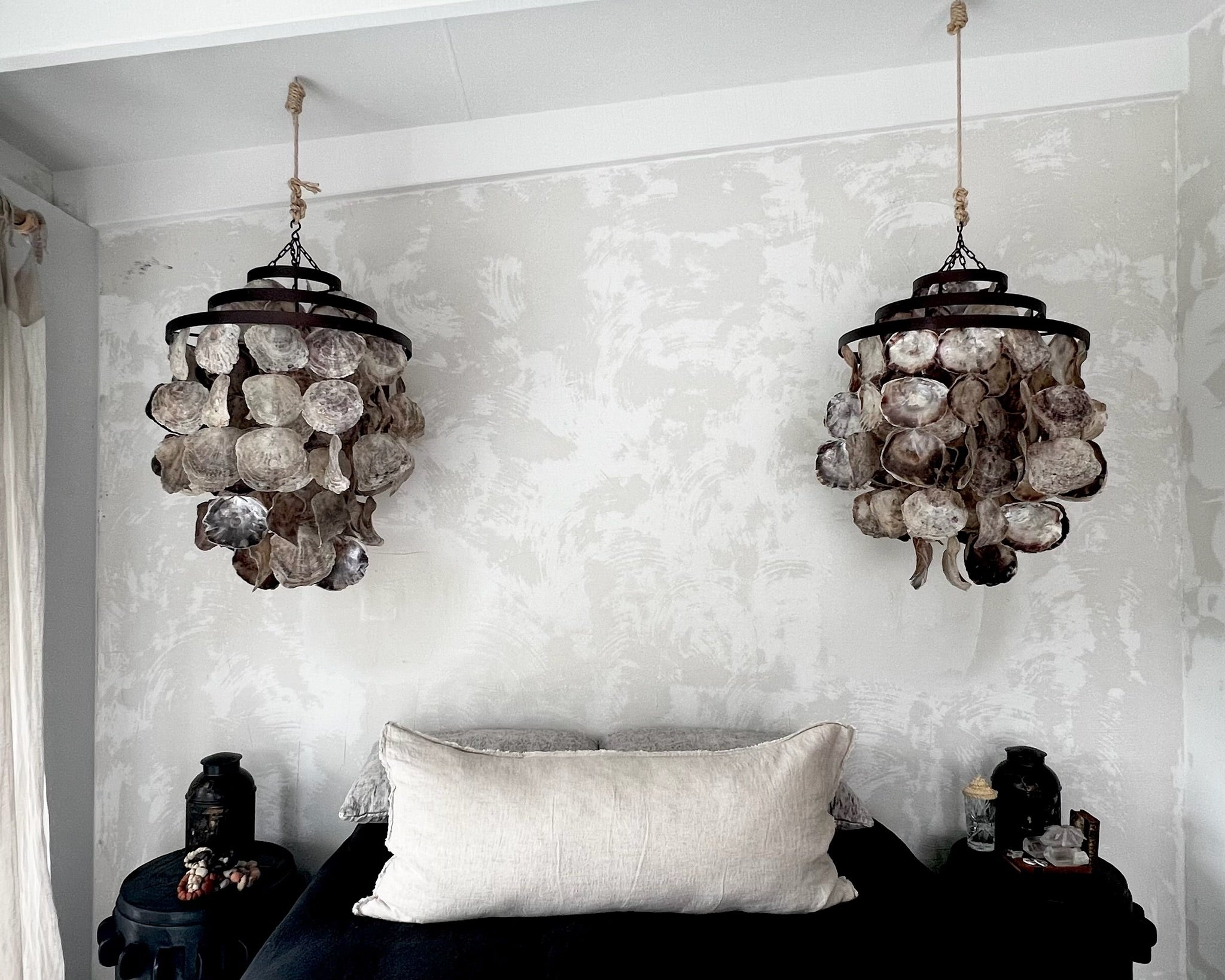 Two decorative chandeliers hanging above a black sofa with a white pillow against a textured white wall.