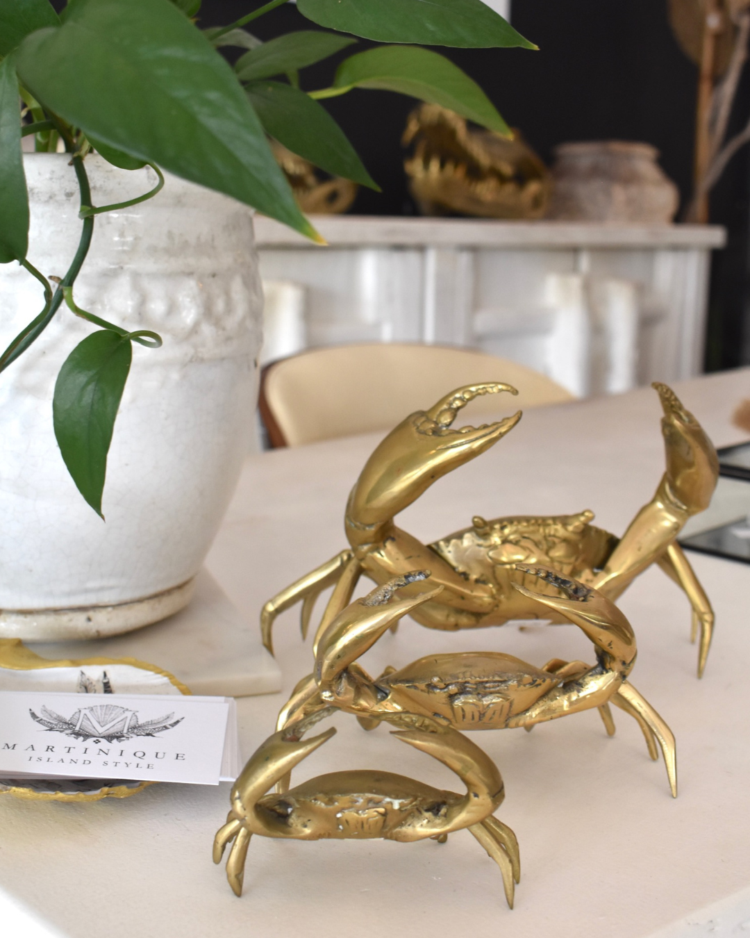 A polished brass decorative crab figure on a wooden surface, with a blurred background featuring stacked white and black round objects.
