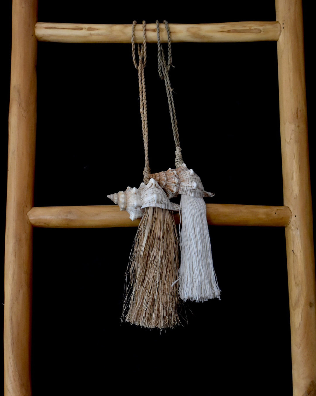 Decorative tassel with shell embellishments hanging from a wooden ladder against a black background