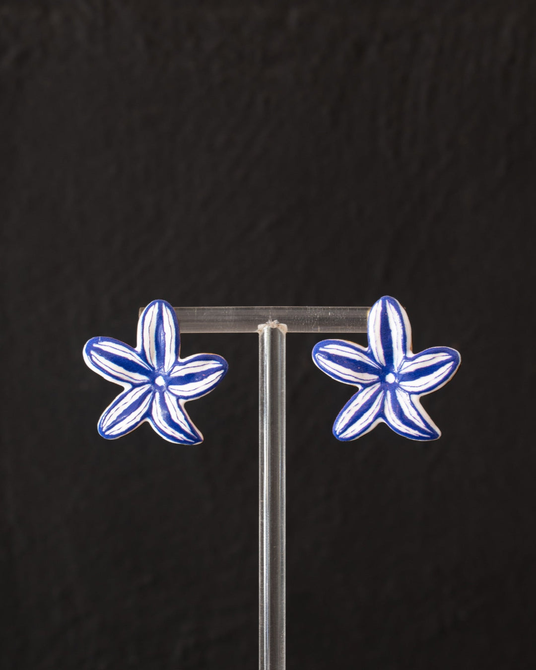 A pair of blue and white flower-shaped stud earrings displayed on a clear acrylic stand against a dark textured background.