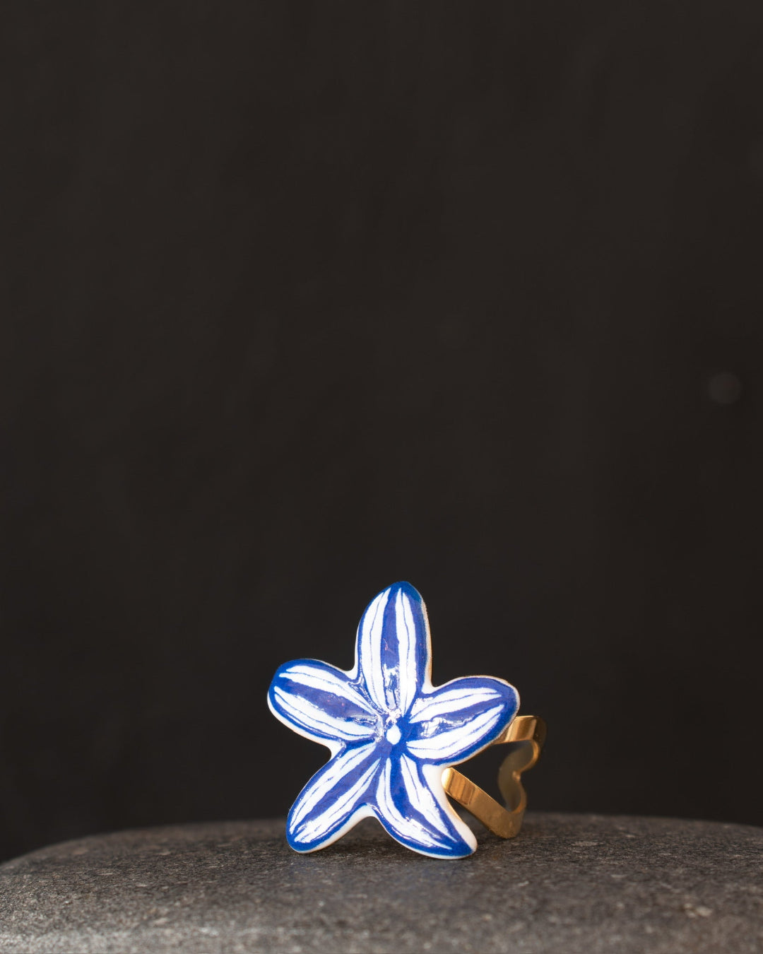 A handmade ring shaped like a five-petal flower, featuring blue and white painted petals with a gold-tone band, resting on a dark gray stone against a dark background.
