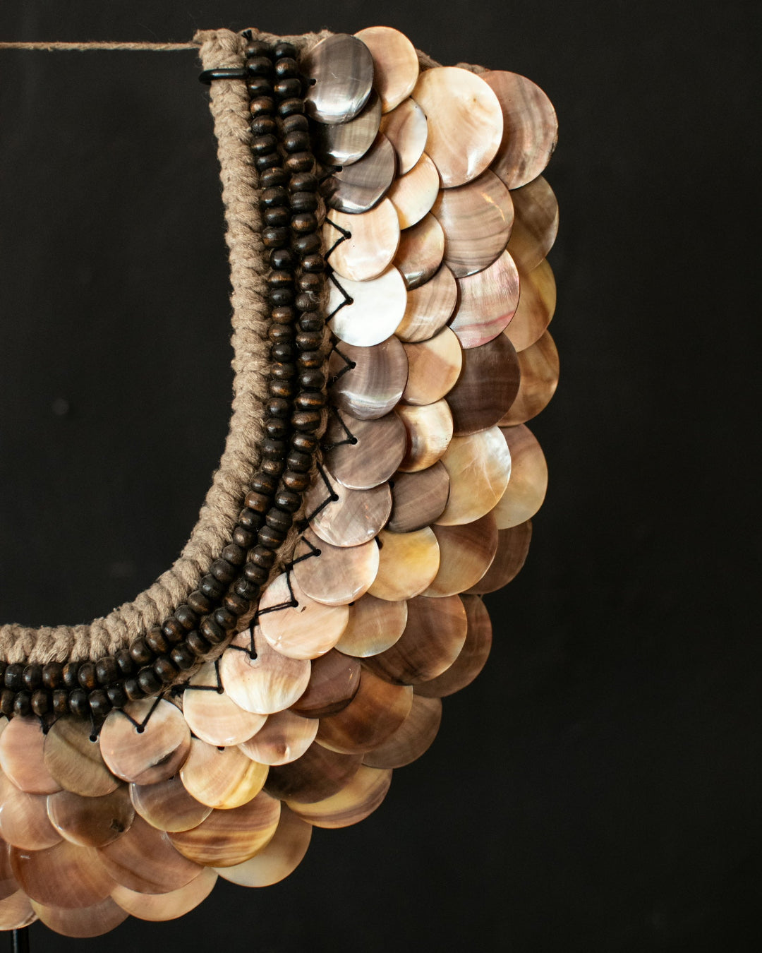 A close-up of a handmade bohemian-style collar necklace featuring a scalloped arrangement of natural wood discs, a thick braided beige cord, and a vertical stripe of dark brown wooden beads, set against a solid black background.