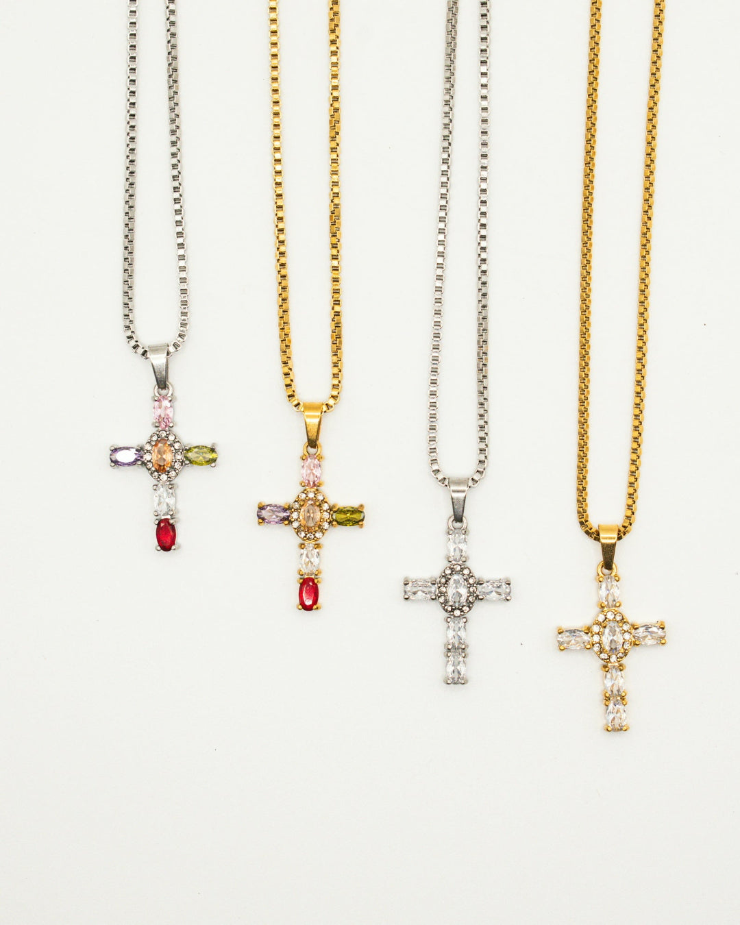 Four cross pendant necklaces on silver and gold chains, each adorned with clear stones and small colored gem accents, displayed on a clean white background.