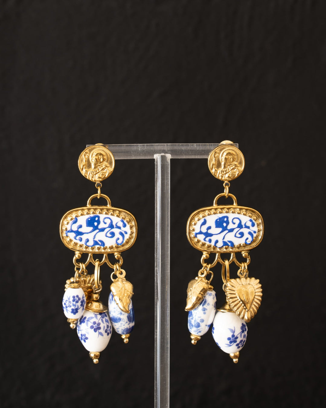 A pair of ornate gold-tone dangle earrings with blue and white porcelain-style floral enamel panels and hanging bead charms, displayed on a clear stand against a dark textured background.
