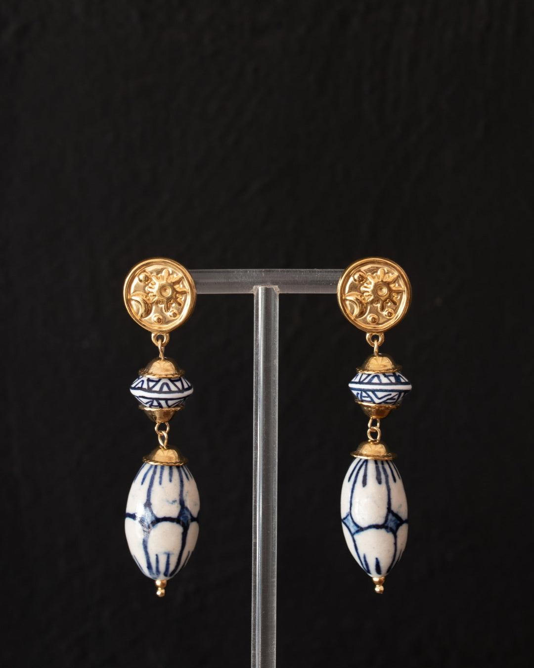 A pair of gold and blue-and-white patterned drop earrings displayed on a clear acrylic stand against a dark textured background.