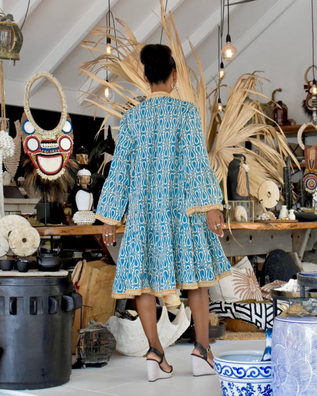 Woman in a blue patterned dress standing in an eclectic interior space with decorative items.