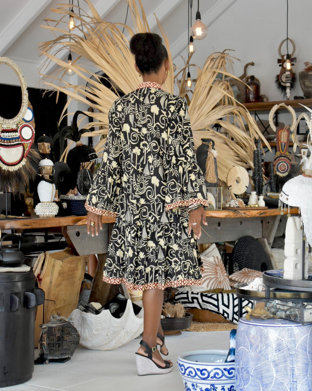Woman in a patterned dress standing in an eclectic interior space with decorative items and plants.