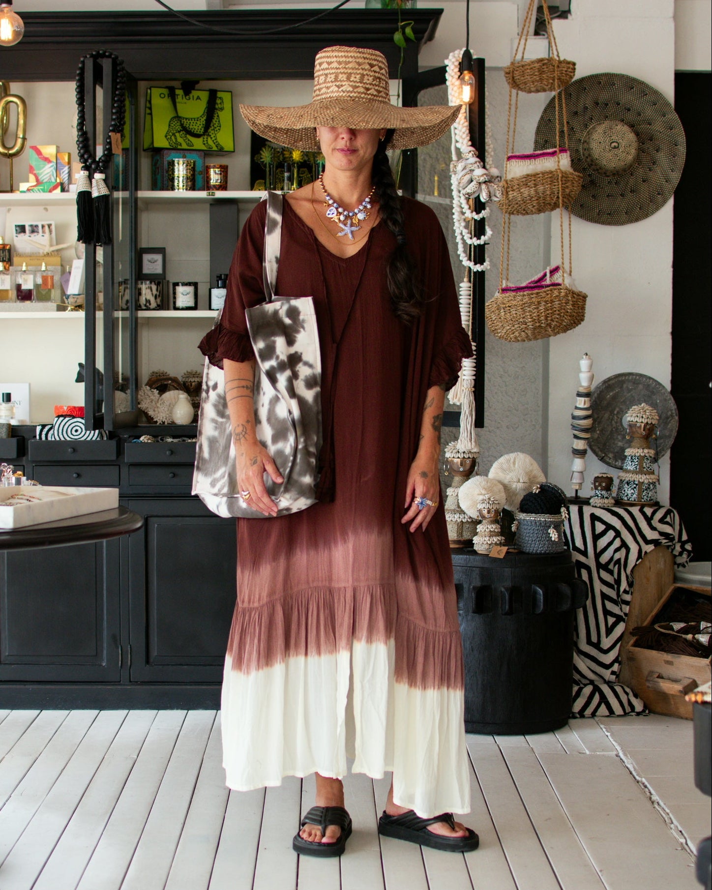 A woman wearing a wide-brim straw hat and a brown and cream tie-dye maxi dress stands in a bohemian-style shop, holding a large patterned tote bag.