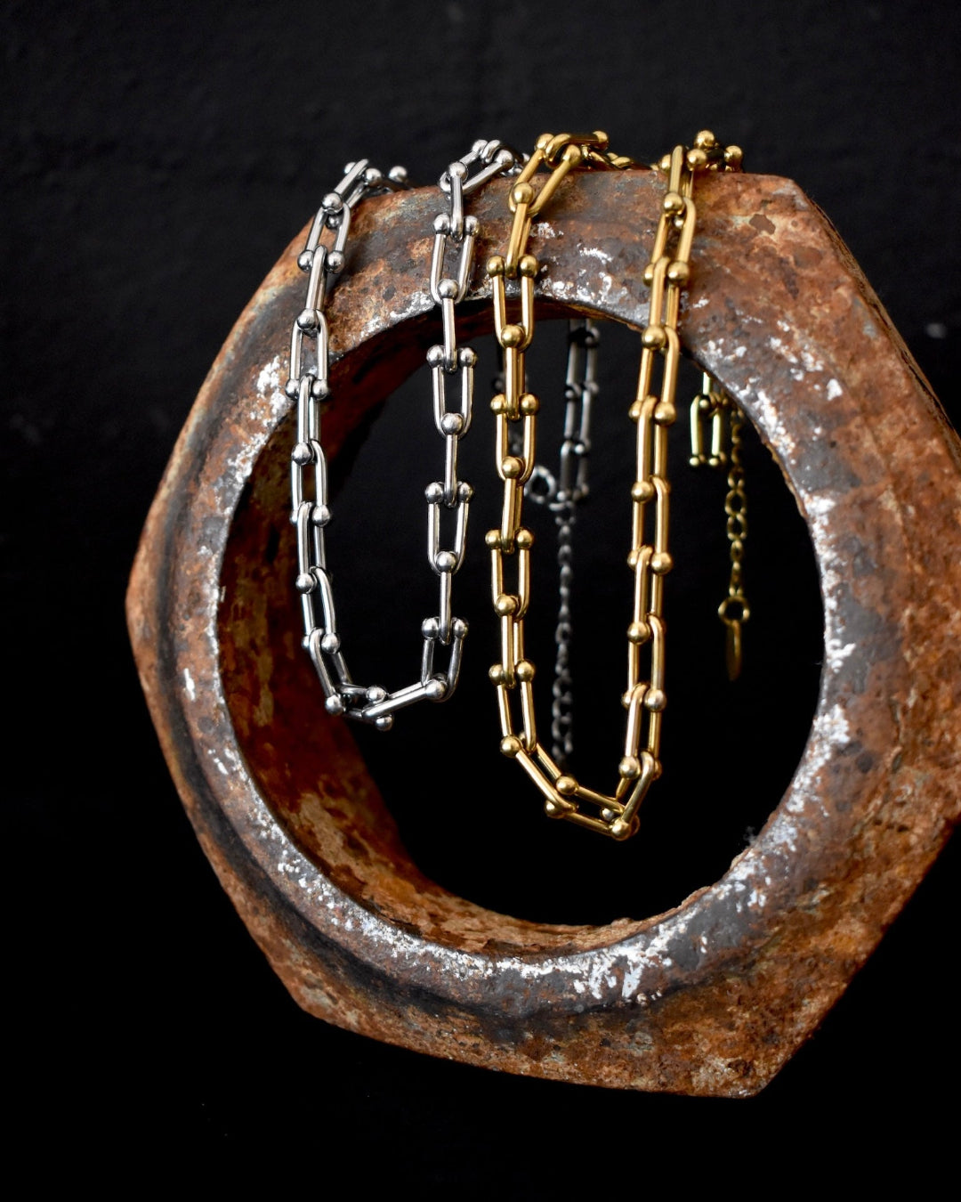 Two chain necklaces, one silver and one gold, draped over a rusted metal hex nut on a dark background.