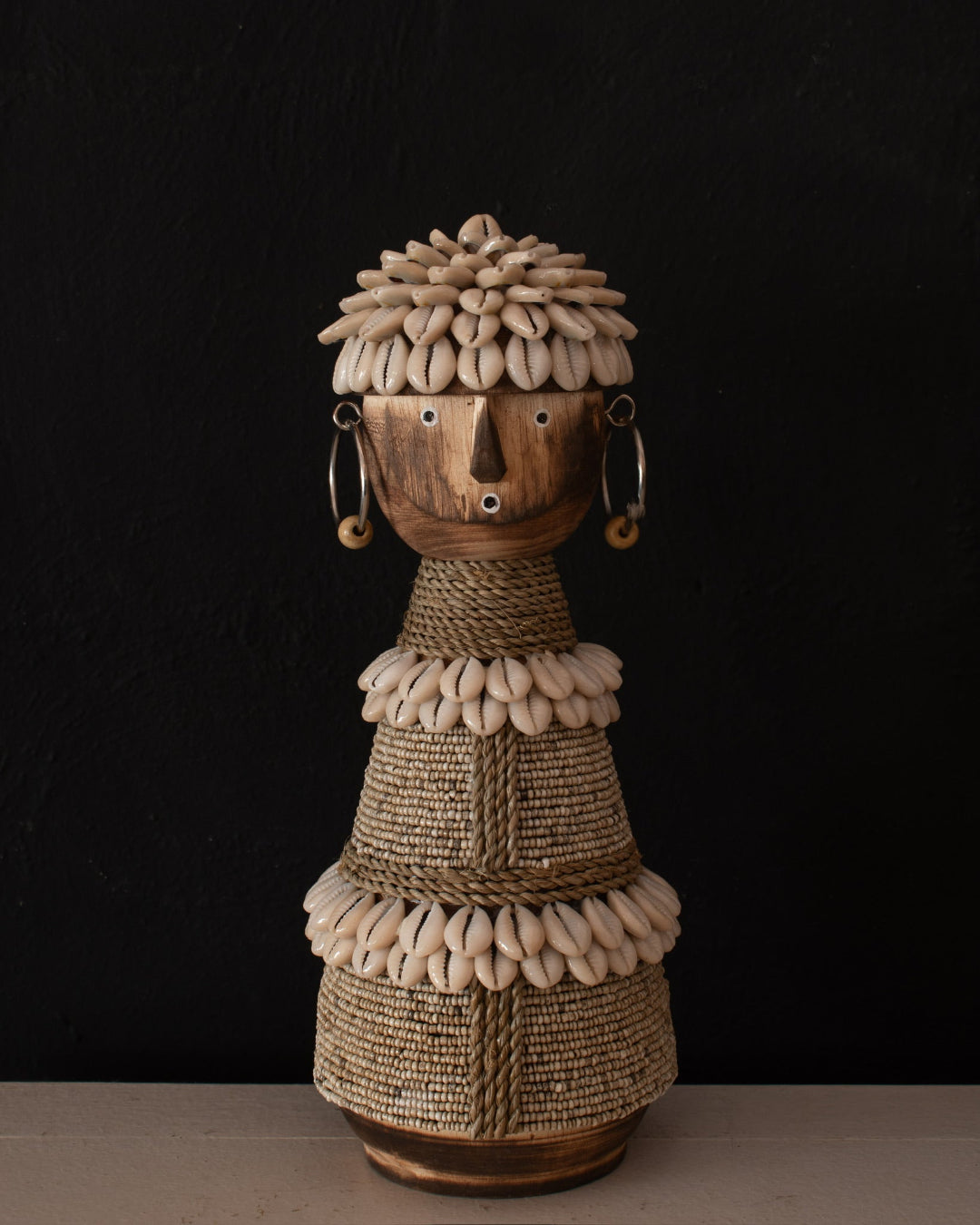 A handmade African-style wooden doll sculpture with a carved face, shell hair, rope details, and black-and-white beaded patterns, set against a dark textured background.