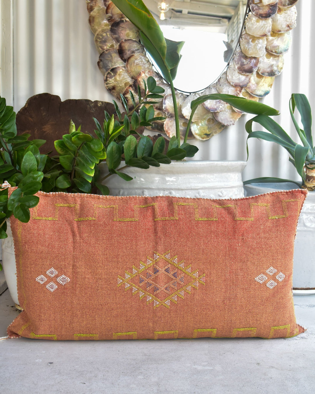 Decorative orange pillow with geometric patterns on a surface with plants and a mirror in the background.
