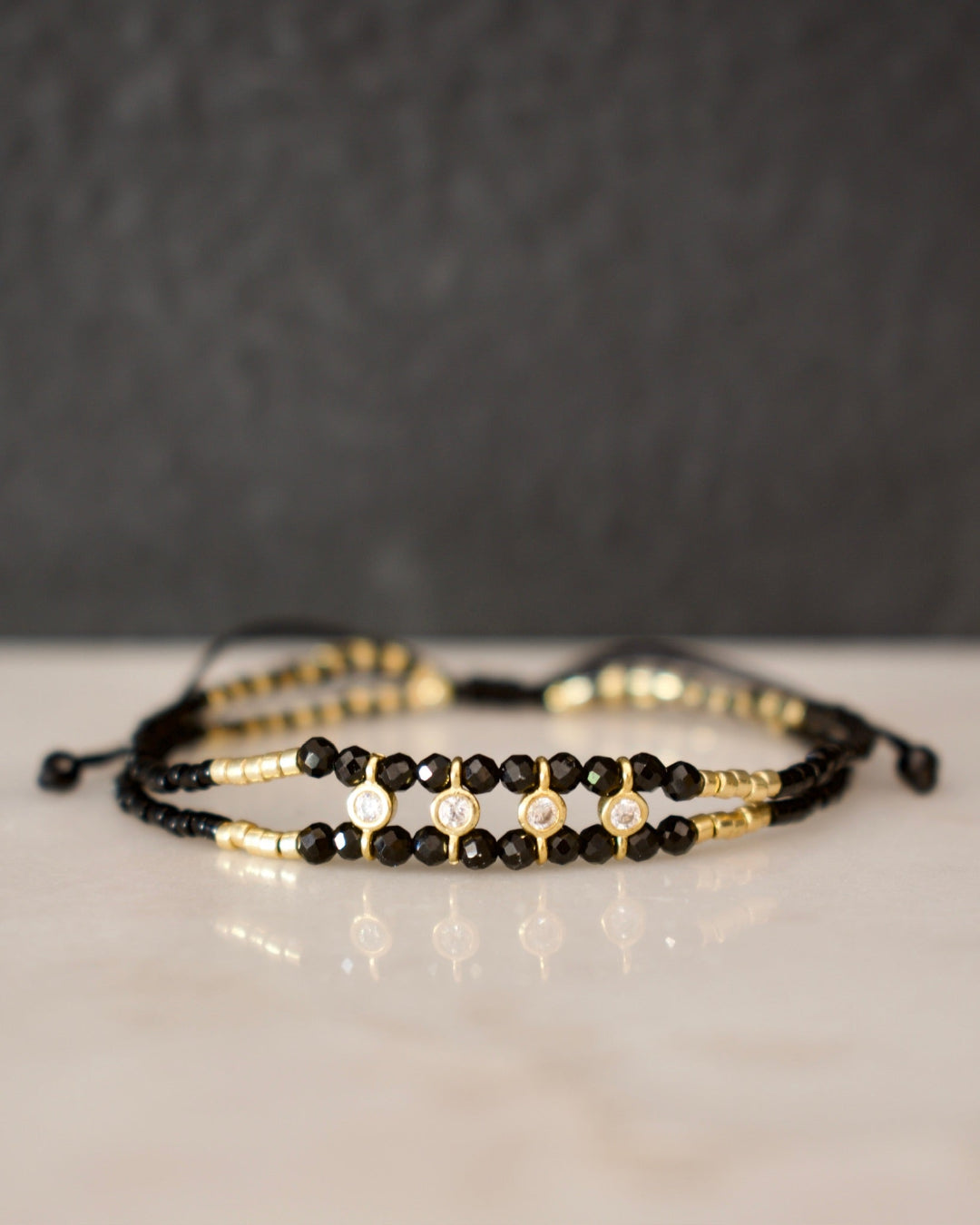 Two beaded bracelets, one black and one gold, on a reflective surface with a dark background.