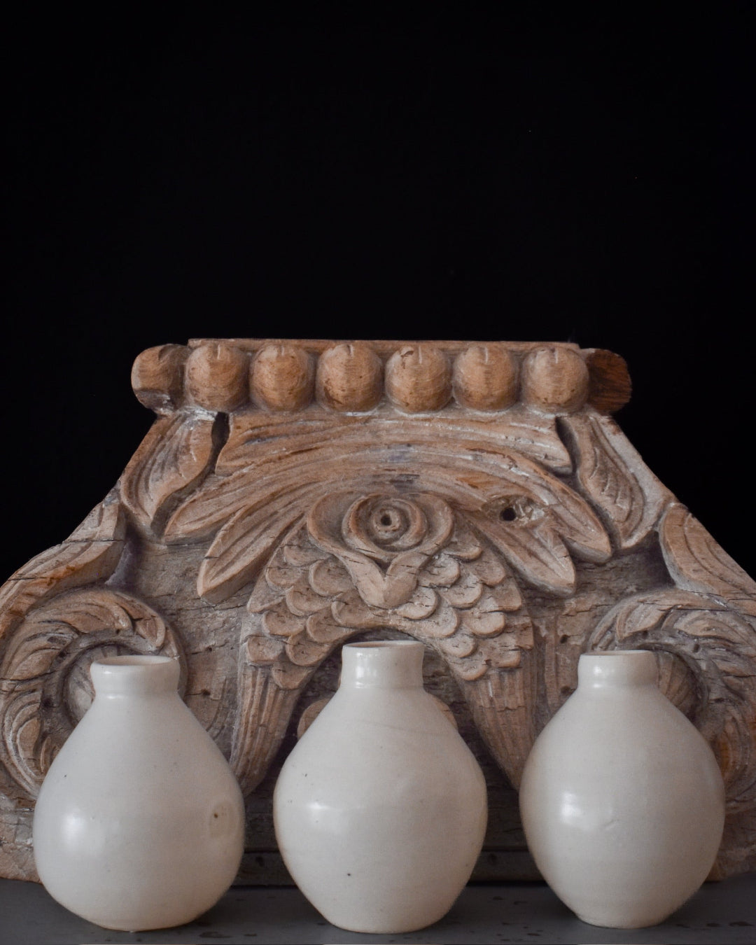 Three white ceramic bottles in front of a wooden carvings on a black background