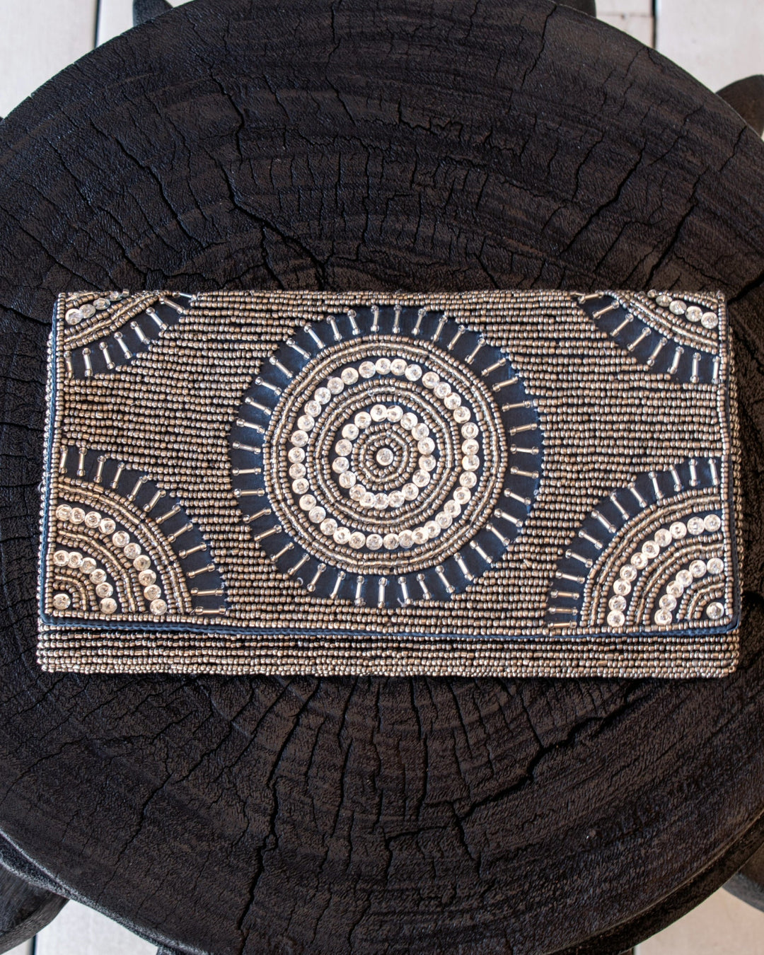 A rectangular beaded clutch purse with a Southwestern-style circular mandala pattern in beige, white, and dark blue, displayed on a dark wood log background.