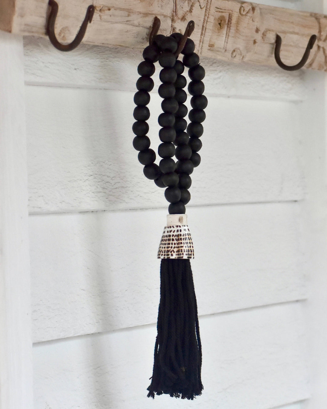 Black beaded tassel hanging on a wooden hook against a white wooden panel background