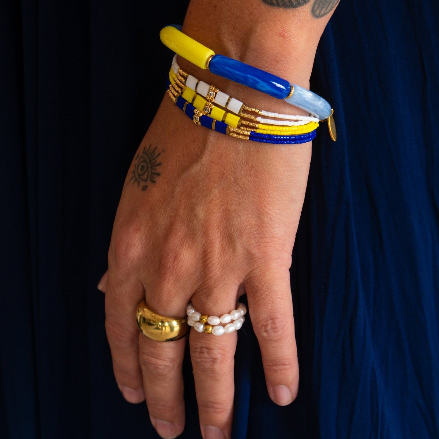 A close-up of a person's hand and forearm wearing multiple bracelets and rings against a dark navy blue fabric background. The bracelets include a yellow and blue bangle, a beaded yellow and blue bracelet, and a white beaded bracelet, while the hand also wears gold and pearl rings.