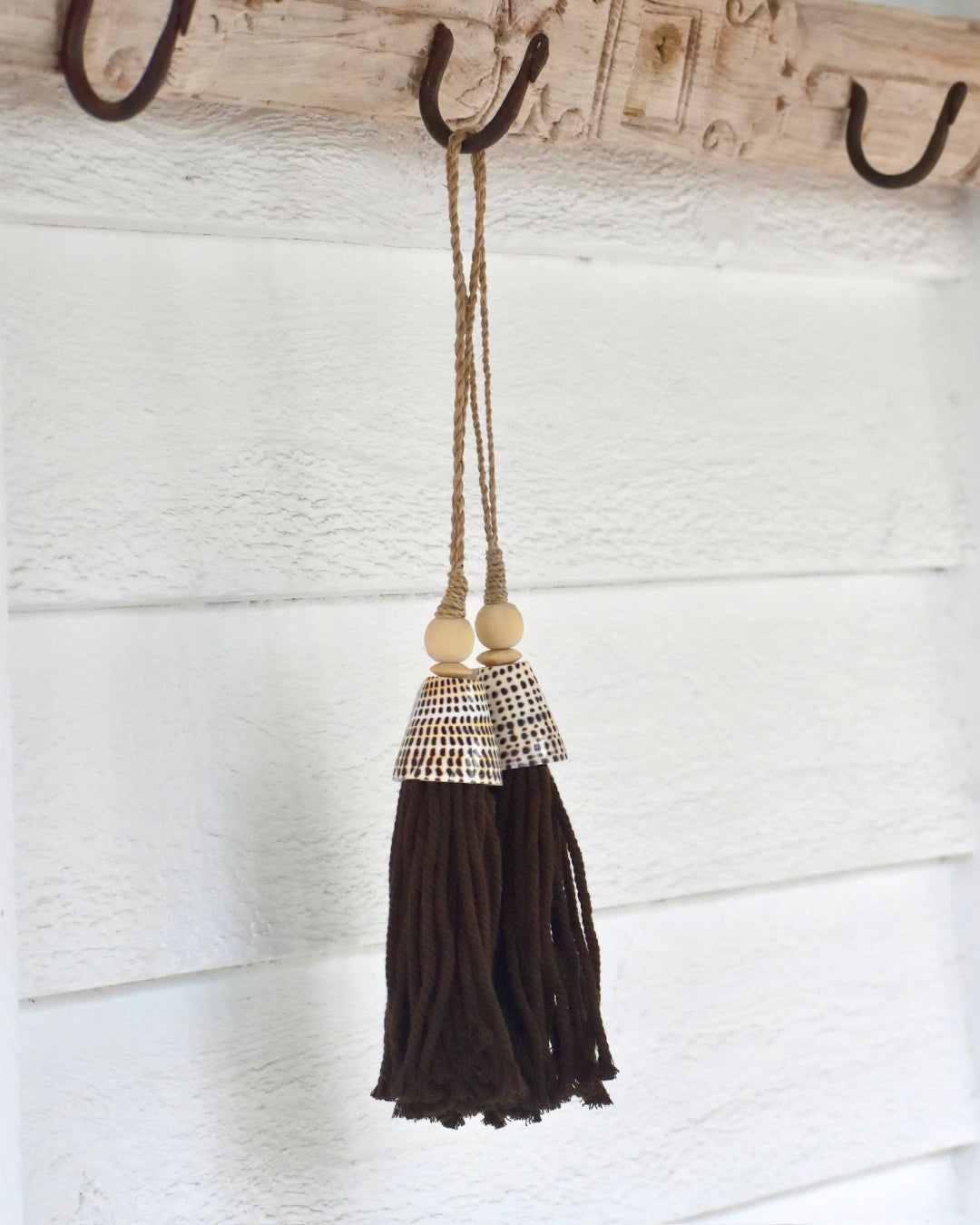 Brown tassel with decorative bead hanging on a wooden hook against a white textured wall.