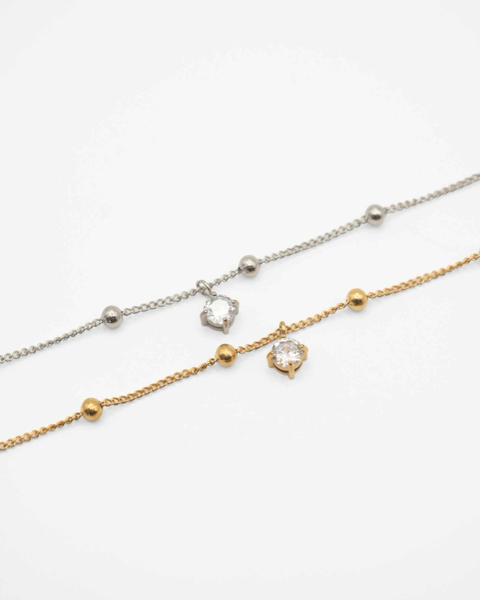 Two delicate chain anklets laid diagonally on a white background. One bracelet is silver-toned with small metallic beads and a clear gemstone pendant.