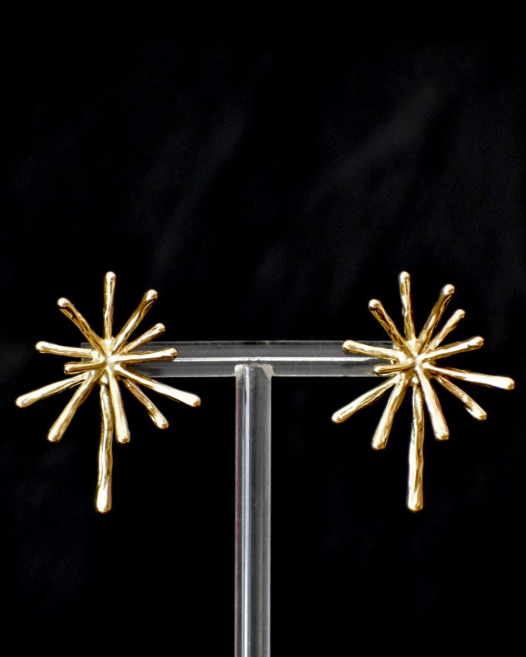 Gold starburst earrings on a clear stand against a black background