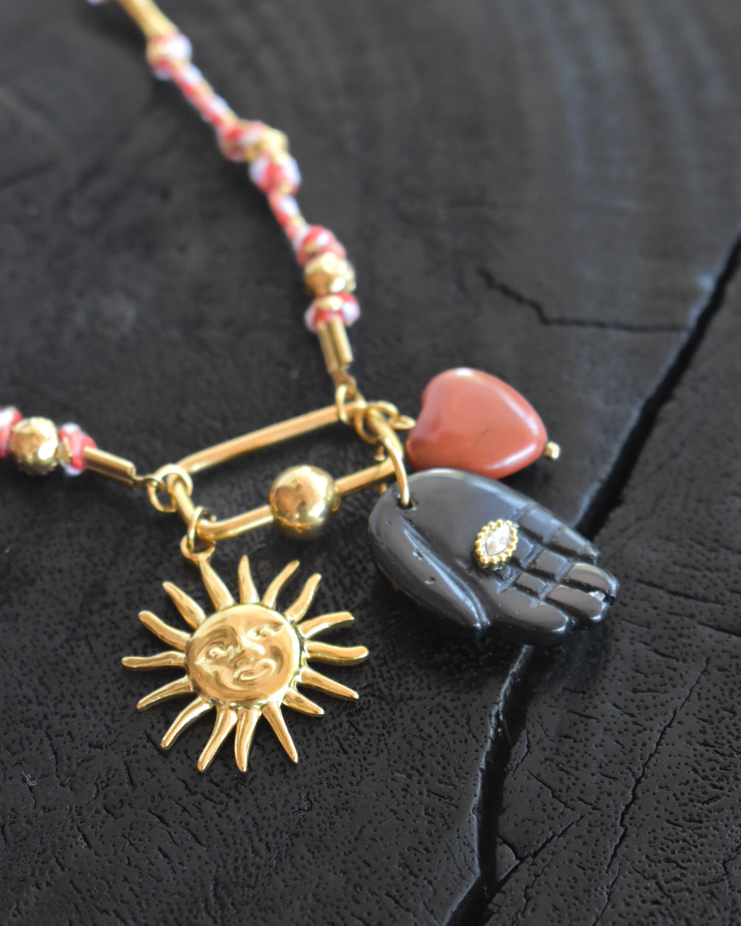 Necklace with sun pendant, heart-shaped red stone, and black hand charm on a dark textured background