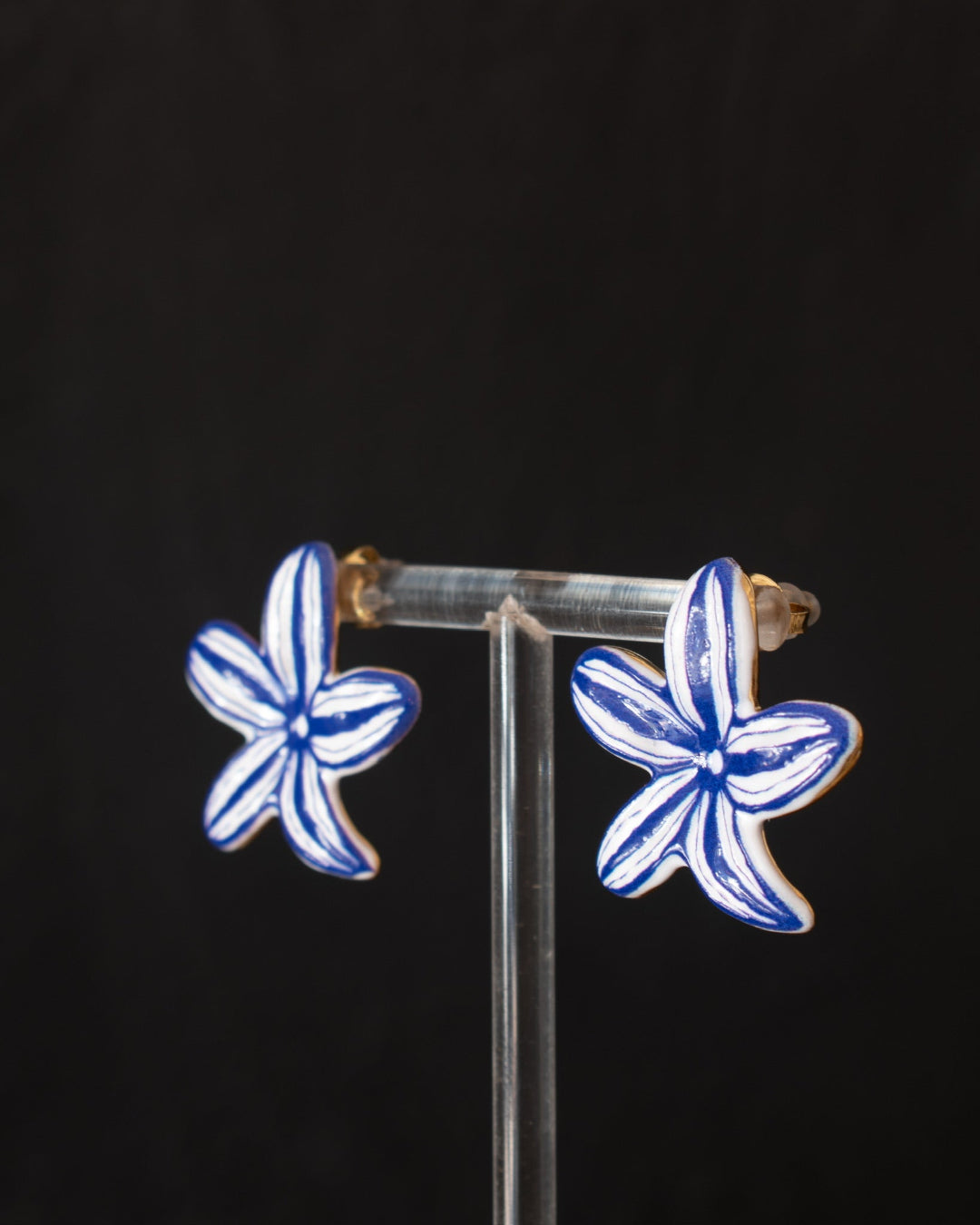 A pair of small flower-shaped earrings with blue and white painted petals, displayed on a clear acrylic stand against a black background.