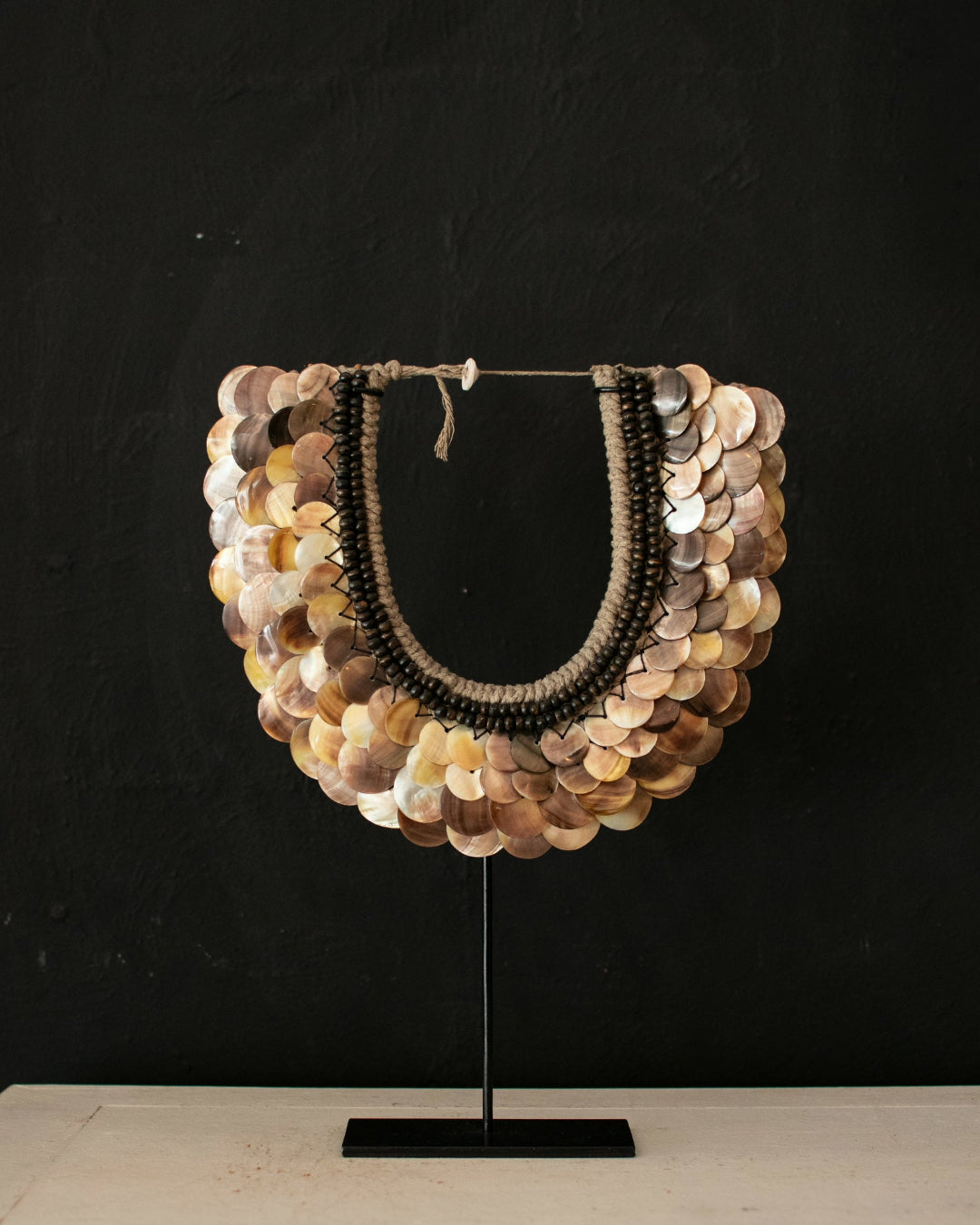 A statement necklace made of layered metallic coin-like discs in gold, bronze, and silver tones, with two dark beaded strands hanging in the center, displayed on a black stand against a dark textured wall.
