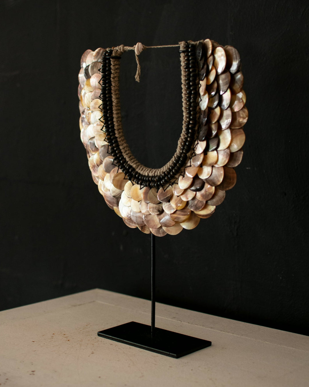 A statement necklace made of overlapping mother-of-pearl shell scales in beige, cream, and brown tones, with dark brown beaded inner strands, displayed on a black stand against a dark background.