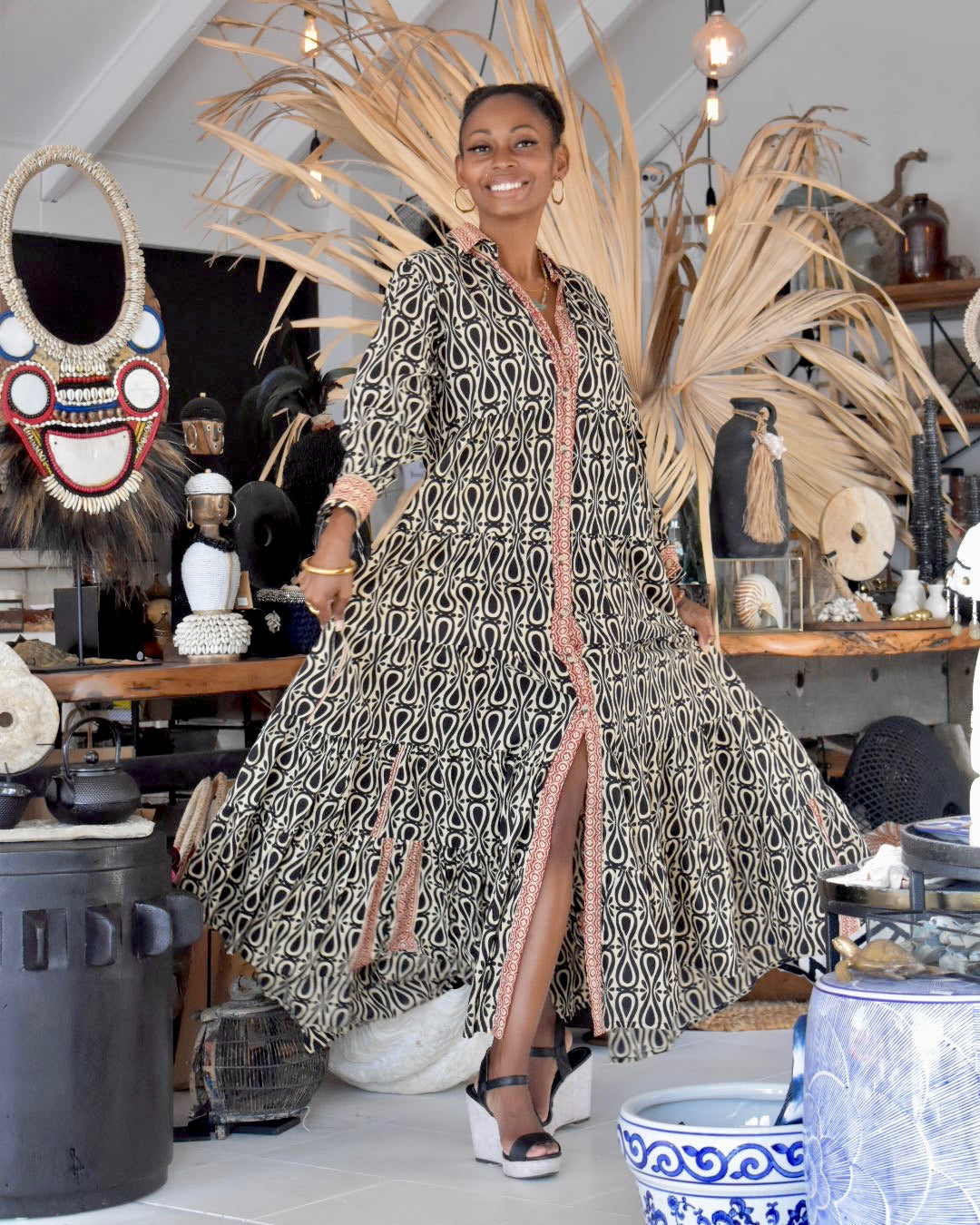 Woman in a patterned dress standing in a decorative interior setting with plants and furniture.