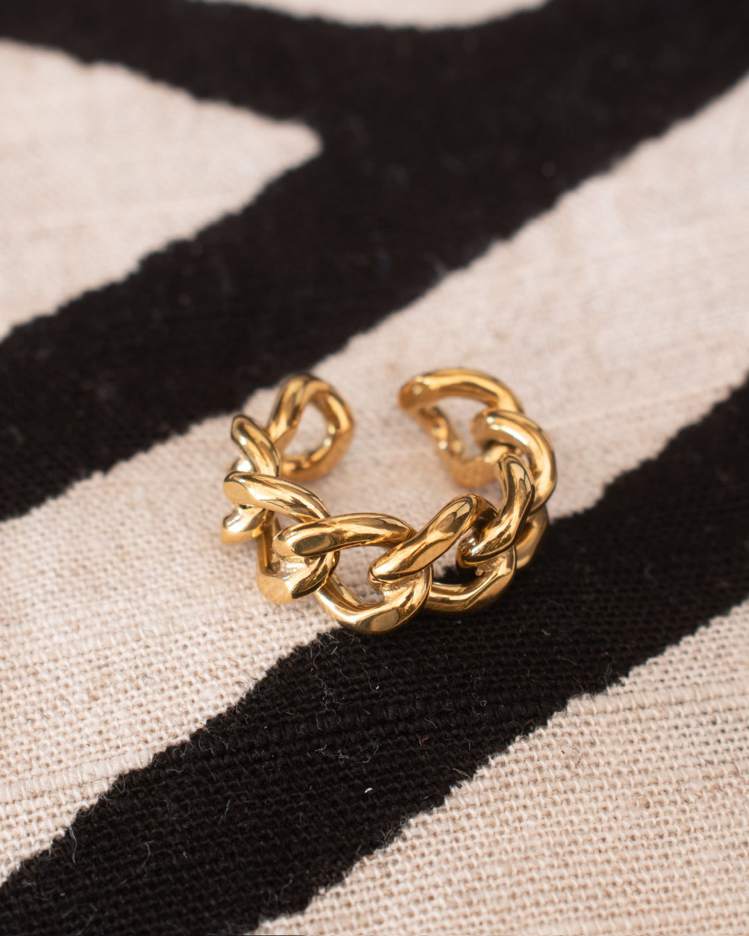 A chunky gold chain ring with a twisted, knotted link design, resting on a black and beige striped knit fabric background.