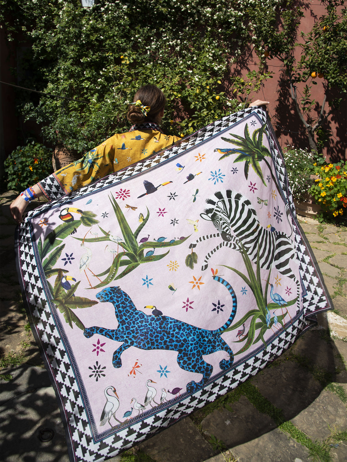 A person outdoors holds up a large square textile featuring a colorful illustrated scene with a blue leopard, zebra, birds, flowers, and a black-and-white patterned border. The background shows greenery and a stone walkway.
