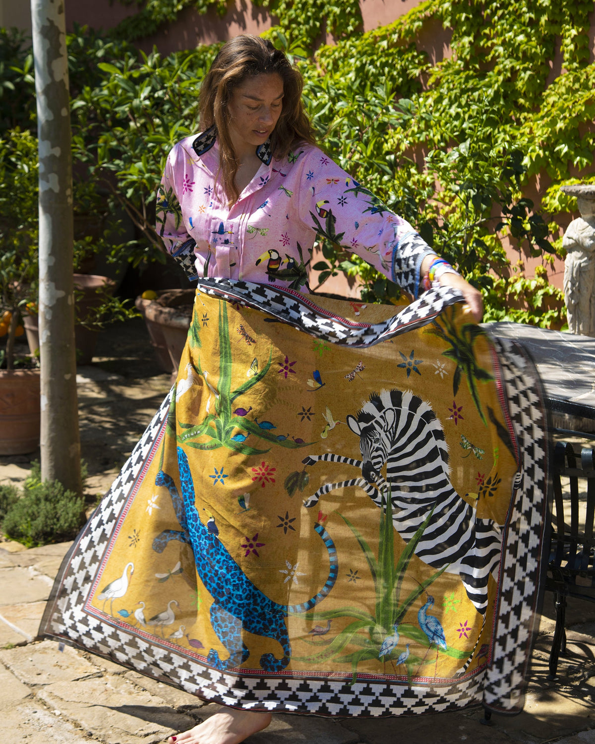 A woman outdoors holds up a large square scarf or wrap with a bold zebra and leopard print on a mustard yellow background, featuring a black-and-white checkered border. She wears a pink patterned shirt, with green foliage and patio furniture in the background.