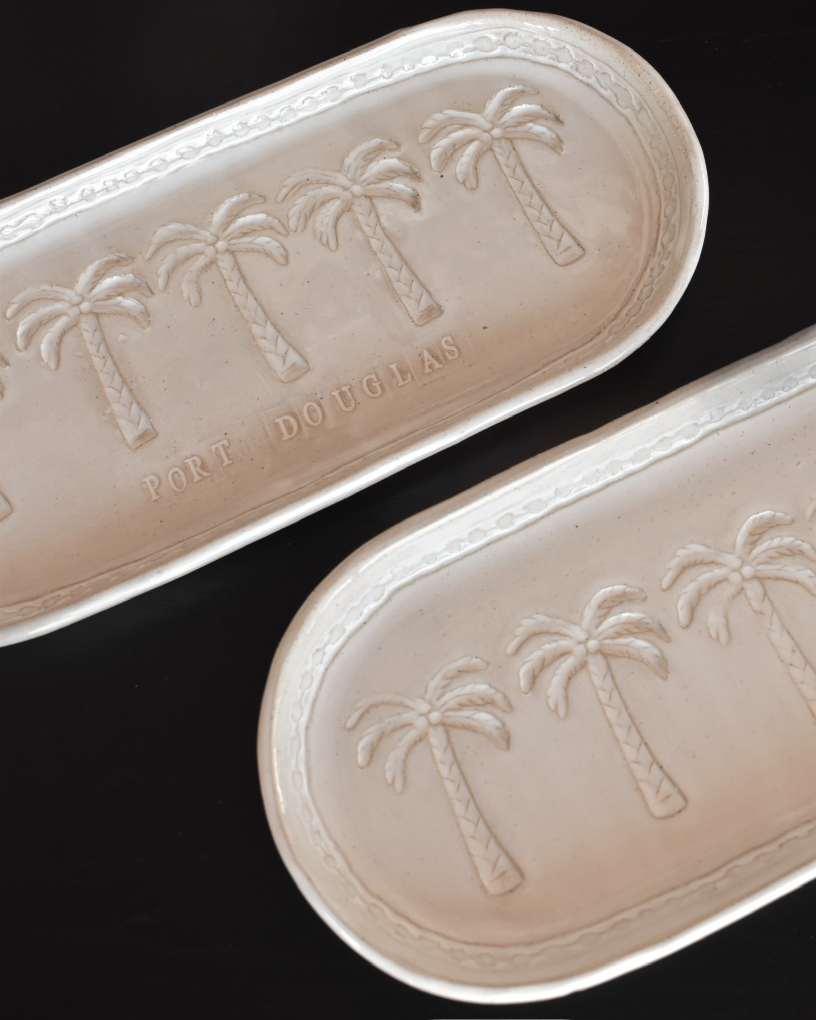 Two beige handmade ceramic trays with palm tree imprint on a black background