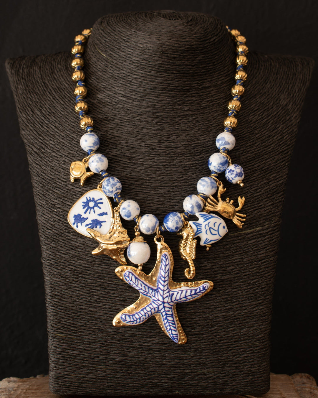 A gold-tone statement necklace displayed on a black textured mannequin bust. The necklace features blue and white patterned beads, a blue and white ceramic seashell charm, a gold seahorse, a gold crab, and a large blue and white enamel starfish pendant.