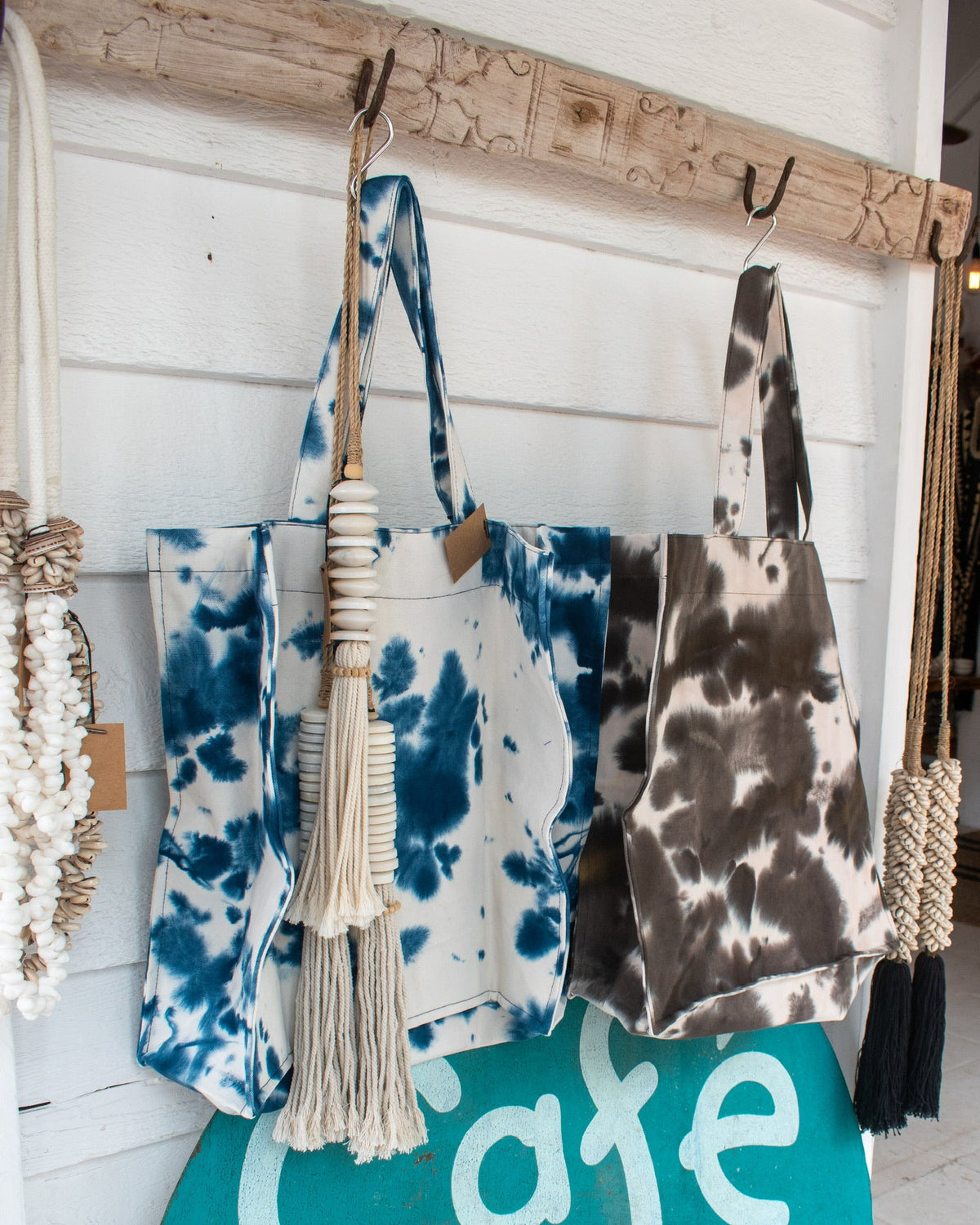 Two fabric tote bags hanging from a rustic wooden rack on a white shiplap wall. One bag is blue and white tie-dye with a cream tassel, and the other is brown and white tie-dye.