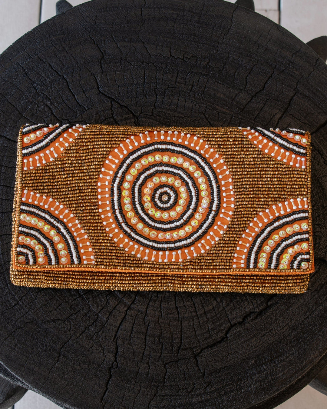 A rectangular beaded clutch bag with an Australian Aboriginal-inspired circular dot pattern in orange, white, black, and brown, displayed on a dark textured tree stump.