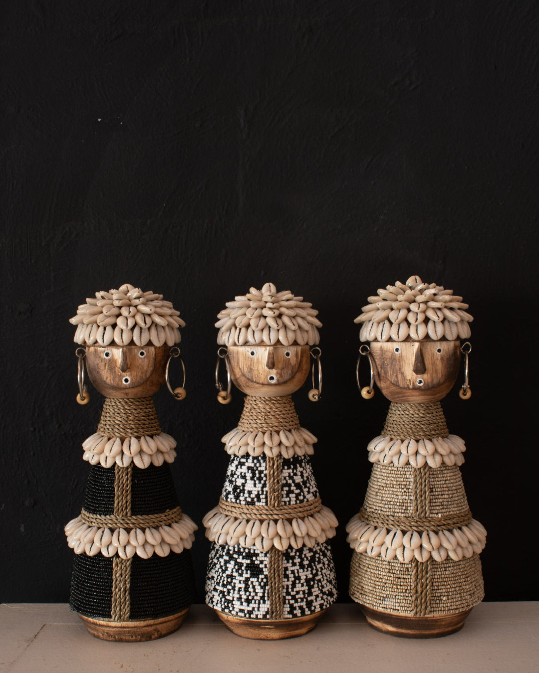 Three handmade African-style wooden doll sculptures decorated with cowrie shells, raffia, and black thread, displayed against a dark textured background.