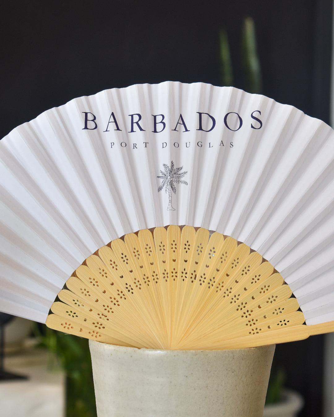 White folding fan with 'Barbados Port Douglas' branding on a blurred background
