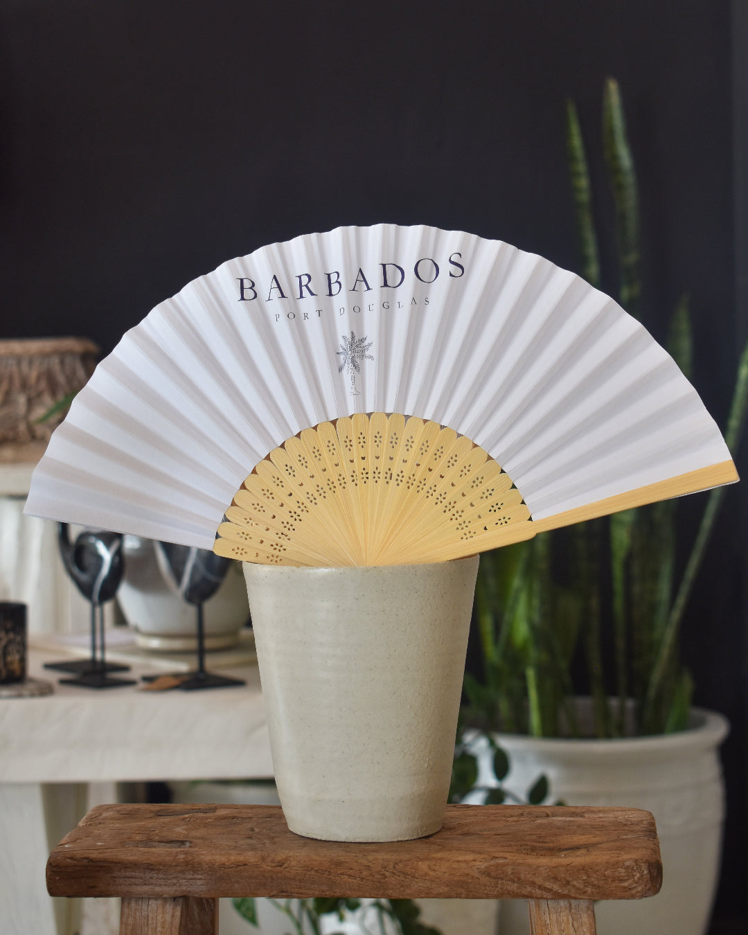 Handheld fan with 'Barbados' branding on a wooden stool with a dark background