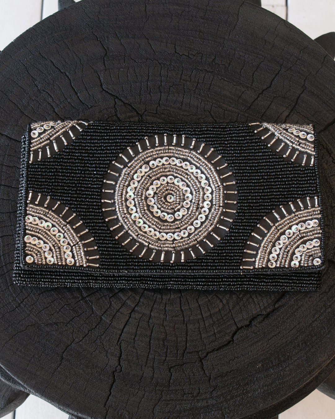 A black beaded clutch purse with intricate circular and geometric patterns in white, beige, and silver, displayed on a dark wood log background.