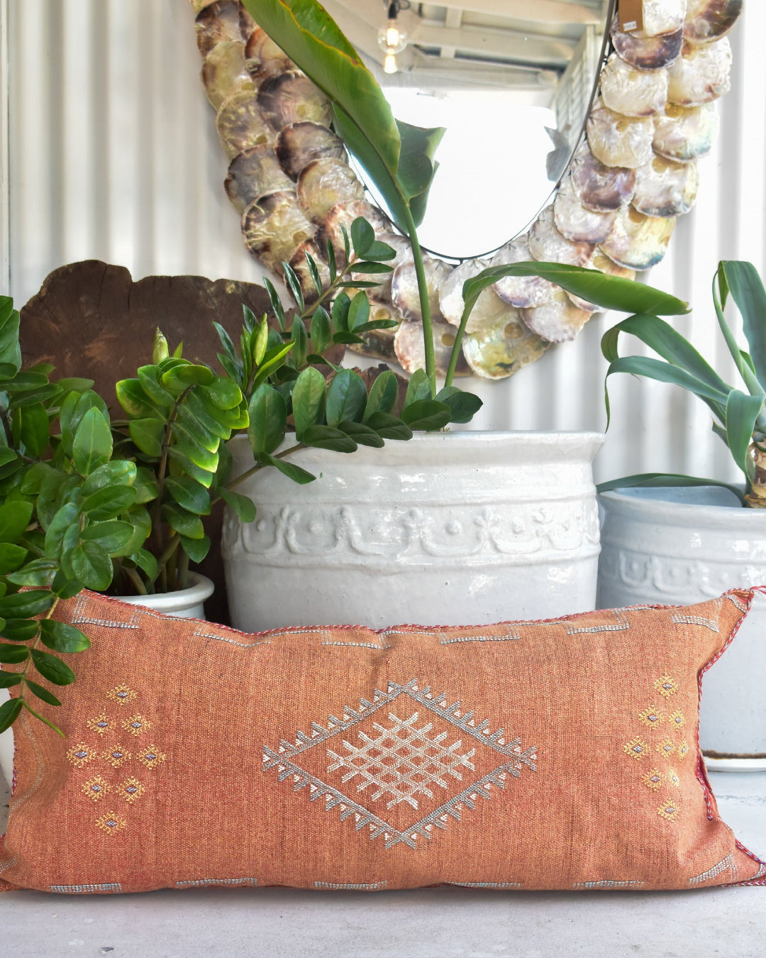 Decorative pillow with intricate patterns on a surface with plants and a mirror in the background.