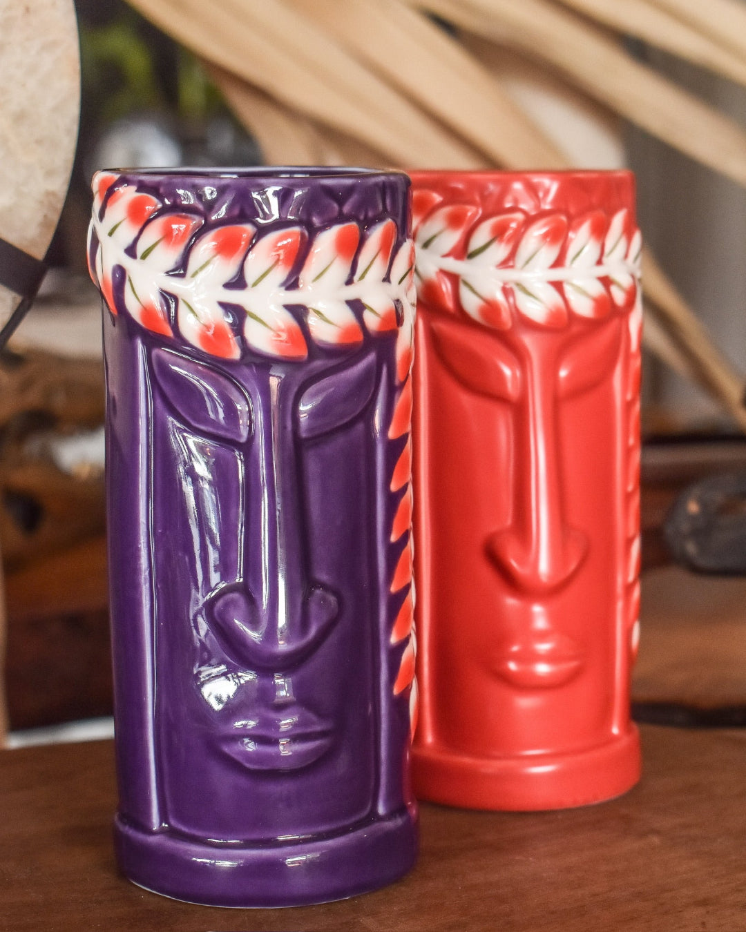 Two tiki mugs, one purple and one red, with face designs on a wooden surface.